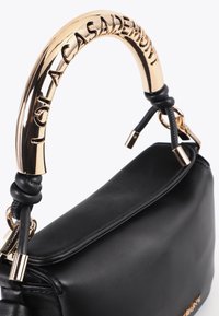 Zwart leren handtas met gebogen gouden handvat gegraveerd met "Lola Casademunt" en zwarte leren knoopdetails.