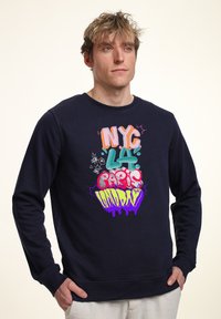 Felpa navy con testo in stile graffiti colorato: "NYC," "LA," "Parigi," e "Londra," con una varietà di colori vivaci e accenti giocosi.