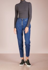 Person in einem grauen langärmligen Rollkragenpullover, hochgeschnittenen blauen Jeans mit Reißverschluss vorne und dunkelgrünen Sneakern mit weißen Sohlen.