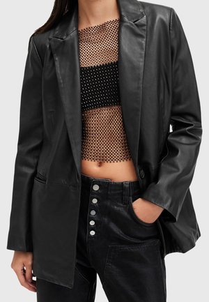 Mujer con una blazer de cuero negra sobre un crop top negro de red y pantalones negros de talle alto con botones plateados, una mano en el bolsillo.