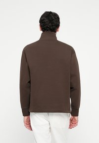 Man sett bakifrån iförd en brun höghalssweater och vita byxor mot en ren vit bakgrund.