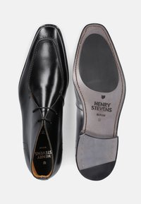 Scarpe eleganti in pelle nera con punta affusolata, lacci decorativi e una finitura liscia. La suola presenta gomma grigia con dettagli di marchio e borchie.