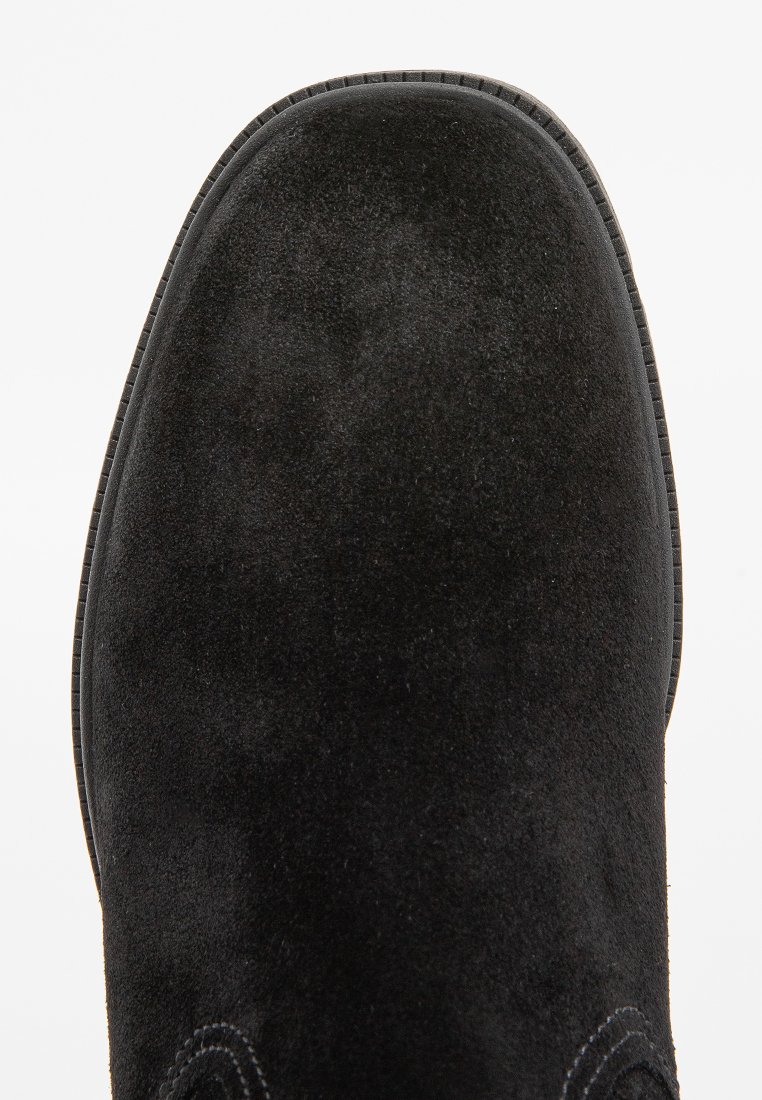 Bottine en daim noir avec le bout visible et des coutures autour de la semelle sur un fond blanc.