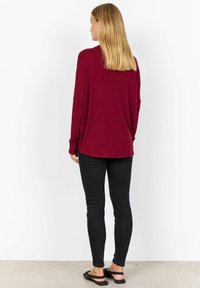 Langarm burgunder top met een ontspannen pasvorm, zachte textuur en iets gebogen zoom, gecombineerd met zwarte skinny jeans en platte sandalen.