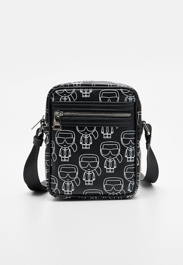 Bolso bandolera negro con figuras de dibujos animados blancas impresas. Cuenta con un bolsillo delantero con cremallera, correa ajustable y material suave y duradero.