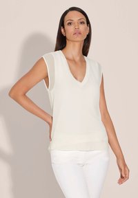 Crème mouwloze top met een V-hals, gemaakt van lichtgewicht stof. Beschikt over een gladde textuur en een relaxte pasvorm, gecombineerd met witte jeans.