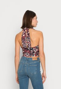 Top halter multicolor com um corte profundo nas costas, apresentando um padrão abstrato em espiral nas cores vermelho, preto e creme, amarrado no pescoço. Combinado com jeans azuis.