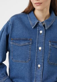 Niebieska koszula dżinsowa z zapięciem na guziki, dwoma kieszeniami na piersiach i klasycznym kołnierzykiem. Szwy są widoczne w kontrastującym żółtym kolorze.