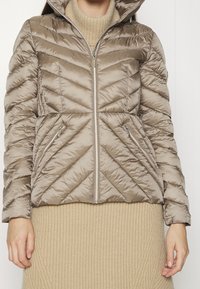 Person trägt eine taupefarbene gesteppte Pufferjacke mit Reißverschluss und zwei Reißverschlusstaschen über einem beigen Strickkleid.