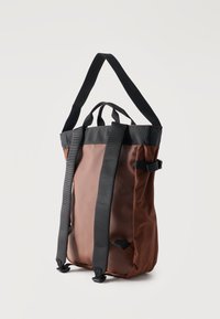 Καφέ και μαύρο σακίδιο με επενδεδυμένους ιμάντες, λαβή στο πάνω μέρος και πλαϊνό κούμπωμα, εμφανίζεται κρεμασμένο σε απλό λευκό φόντο.