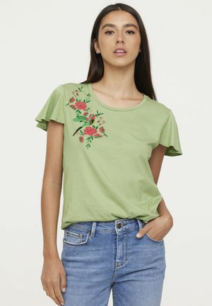 T-shirt en coton vert clair avec des manches courtes à volants, présentant une broderie florale rouge sur le côté gauche. Porté avec un jean bleu.