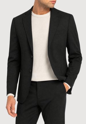 Mann trägt eine schwarze, maßgeschneiderte Anzugjacke und passende Hose mit einem weißen Rundhalspullover darunter, Hand teilweise in der Tasche.