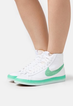 Baskets montantes blanches avec un Swoosh Nike vert texturé et des semelles vertes portées sur des jambes nues sur un fond blanc.