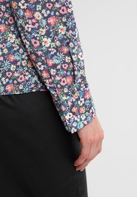 Blouse fleurie avec un fond bleu marine, ornée de fleurs roses, blanches et bleues ; manches longues, poignet boutonné et texture en coton lisse.