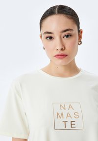Cremefarvet t-shirt med en firkantet grafik i guld folie med teksten "NAMASTE", korte ærmer og en rund halsudskæring.