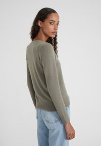 Femme avec de longs cheveux bouclés portant un pull vert et un jean bleu clair, se tournant vers son épaule devant un fond gris uni.