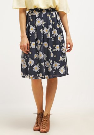 Vrouw draagt een marineblauwe geplooide rok met wit madeliefjespatroon, een geel topje en bruine veterlaarsjes met open teen, staande tegen een witte achtergrond.