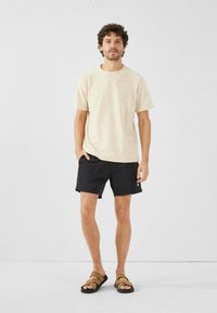 T-shirt en coton beige, shorts noirs, sandales marron, pose debout avec une main dans la poche, léger texturage sur le tissu, fond neutre.