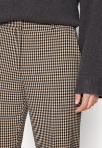 Gros plan d'une personne portant un pantalon à carreaux beige et noir et un pull gris foncé à manches longues, la main reposant près de la cuisse.