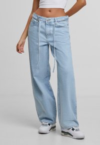 Žena v volných světlomodrých džínách s šňůrkou v pase a bílými teniskami, stojící na světlém jednobarevném pozadí.