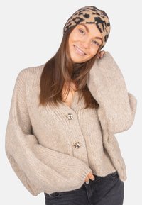 Cardigan en maille beige avec des manches surdimensionnées, orné de grands boutons décoratifs. Associé à un serre-tête à imprimé léopard. Texture douce.