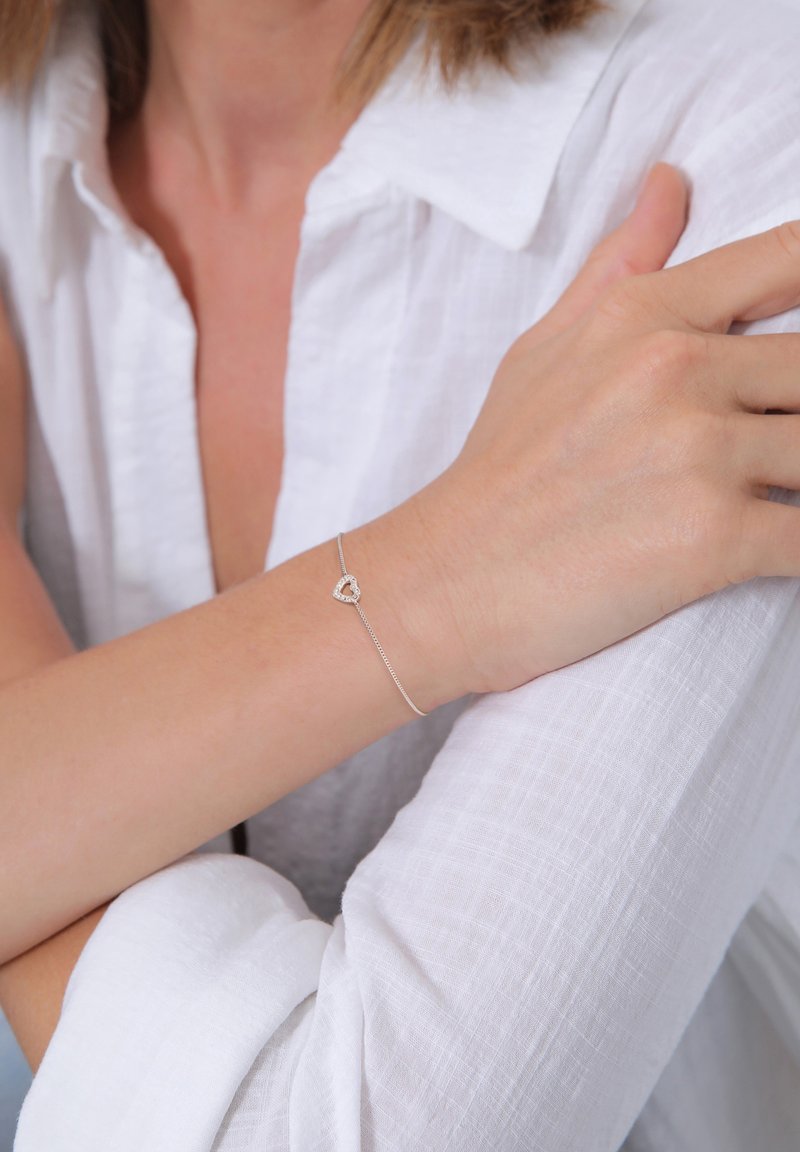 Silverarmband med ett hjärtcharm, som har en delikat kedjedesign. Bar på en handled, med en bakgrund av en texturerad vit tyg.