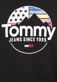 Jersey negro con un gráfico circular que presenta el texto "Tommy" en blanco, montañas, estrellas de la bandera americana y franjas horizontales en azul, amarillo y rosa.