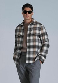 Karierte Flanellhemd in Grau, Braun und Weiß mit zwei Brusttaschen, kombiniert mit einem braunen Rippenpullover und grauen Hosen. Schwarze Sonnenbrille getragen.