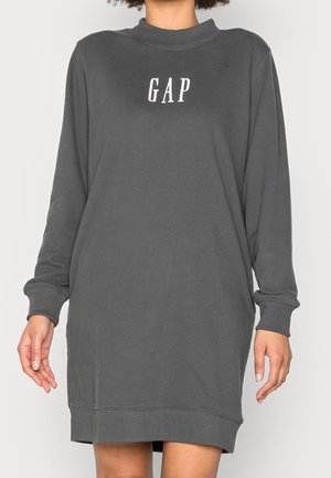 Kobieta w luźnej, długorękawowej sukience typu sweatshirt w ciemnoszarym kolorze z napisem "GAP" na piersi w białych literach.