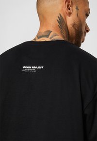 Camiseta de algodão preta com decote arredondado, mangas curtas e impressão em branco nas costas: "DENIM PROJECT 2018 COPENHAGEN."