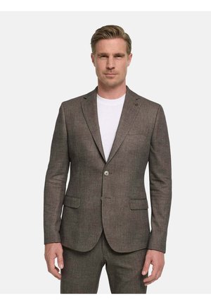 Mann trägt eine braune maßgeschneiderte Anzugjacke und passende Hose über einem weißen Rundhalsausschnitt, steht vor einem schlichten hellen Hintergrund.