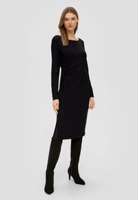 Schwarzes langärmliges Kleid mit quadratischem Ausschnitt, seitlicher Raffung und seitlichen Schlitzen. Getragen mit knielangen schwarzen Stiefeln. Glatter, strukturierter Stoff.