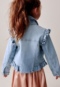 Ragazza con lunghi capelli castani raccolti in una coda di cavallo, indossa una giacca in denim azzurro chiaro con maniche e orlo arricciati, vista di spalle su uno sfondo semplice.
