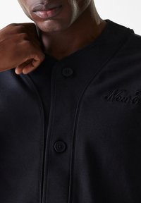 Chemise noire à boutons, avec un col arrondi, une texture lisse et un détail brodé "New&". Les boutons sont grands et sombres.