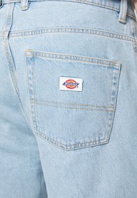 Light blue denim jeans with a back pocket, featuring a white label with a red and yellow logo. Stitching is visible along the seams.