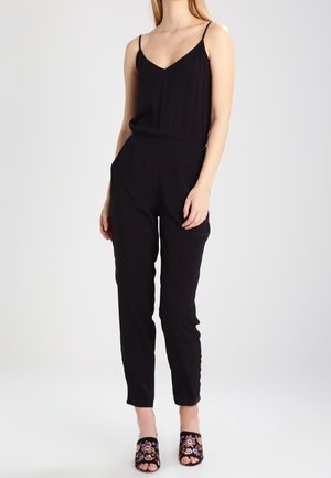 Femme portant une combinaison sans manches noire avec un décolleté en V et une taille élastique, associée à des mules noires à bout ouvert brodées de fleurs.