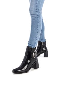 Botines de charol negro con un tacón grueso, puntera afilada y detalle de hebilla plateada, combinados con jeans ajustados de color azul claro.