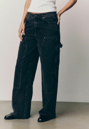 Mustat denim cargo-housut rentoa mitoitusta, joissa on suuret sivutaskut, näkyvät ompeleet ja rispaantunut helma alhaalla.