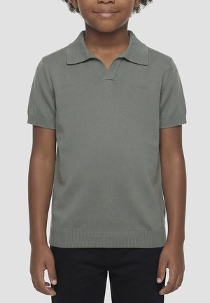 Niño con polo tejido de manga corta y color gris claro, con cuello, de pie sobre un fondo liso.