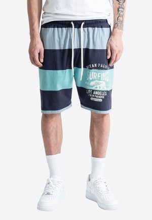 Mand iført marineblå, blågrønne og lyseblå stribede shorts med surfing-tekst, hvide sokker, hvide sneakers og en hvid T-shirt, stående.