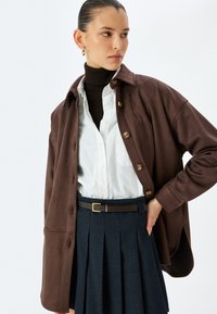 Chaqueta marrón abotonada sobre una blusa blanca, combinada con un cuello de tortuga de canalé. Presenta una falda plisada azul marino y un cinturón marrón con una hebilla dorada.