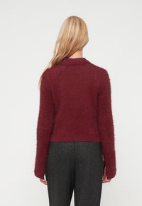 Blond oseba stoji obrniena stran, oblečena v puhasto burgundsko sveter in temno sivo karo hlače pred enobarvno belo háttje.