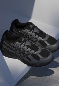 Par sorte og mørkegrå atletik-sneakers med mesh og syntetiske paneler, sorte snørebånd og såler med mønster, placeret på en hvid kantet overflade.