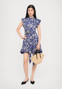 Floral print dress in blue with short sleeves and a flared skirt. Features a tie waist and tassel accents. Small woven handbag in beige.