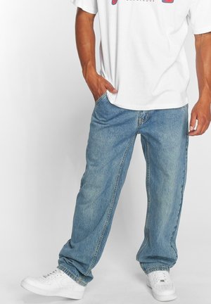 Person wearing loose light blue jeans, white sneakers, and a white T-shirt with red and blue text, standing with one hand in pocket.