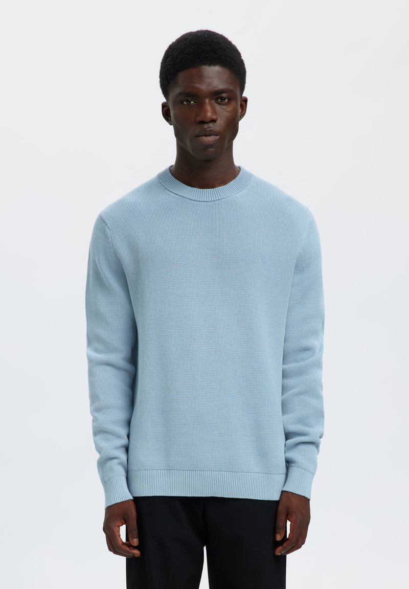 Jeune homme portant un pull en tricot bleu clair et un pantalon noir, se tenant debout avec une expression neutre sur un fond blanc uni.
