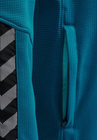 Teal träningjacka med texturerad tyg, en sidoficka med dragkedja och en kontrasterande svart sektion med vit randig zigzag-mönster.