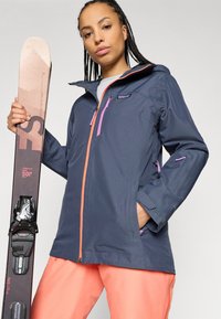 Ναυτικό αδιάβροχο μπουφάν σκι με κουκούλα, ροζ φερμουάρ και ρυθμιζόμενους καρπούς. Μοντέλο που κρατάει σκι φορεί φωτεινά πορτοκαλί παντελόνια σκι.