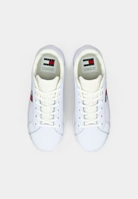 Białe sneakersy o gładkiej skórzanej powierzchni, z sznurówkami, wyściełanym kołnierzem i czerwono-czarnym logo po boku.