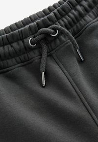 Schwarze Jogginghose mit strukturertem Bund, verstellbarem Kordelzug und metallischen Ösen mit Kunststoffspitzen. Glatte Stoffoberfläche.